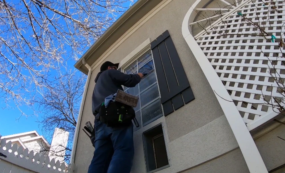 Window cleaning technician on a ladder cleaning an upper-floor window with shutters in San Clemente